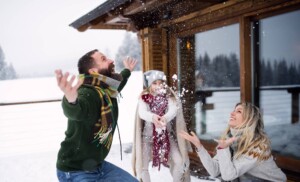 Jeune famille de deux parents et une fillette jouant avec la neige devant un chalet en nature.