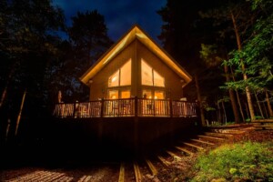 Chalet de bois en forêt illuminé de l'intérieur le soir, avec une grande galerie.