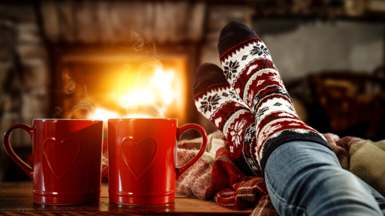 Cozy romantic scene with two steaming red mugs and a couple relaxing by the fireplace, representing romantic cottage getaways in Ontario.